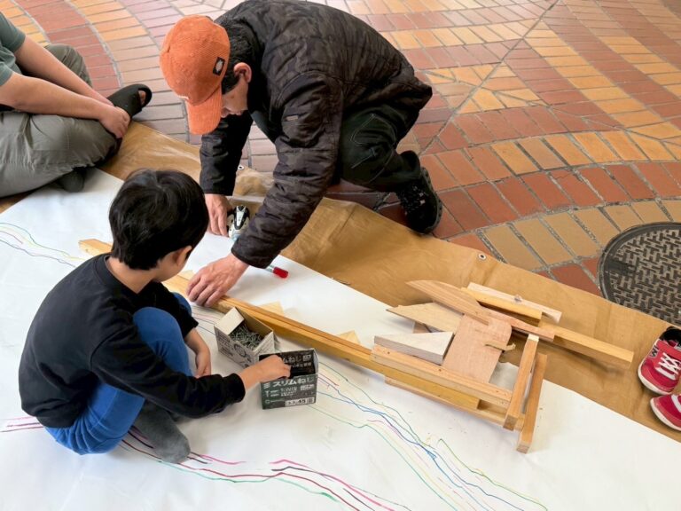 男の子が木で恐竜を作っており、それを手伝うハブラボ講師ケンケン