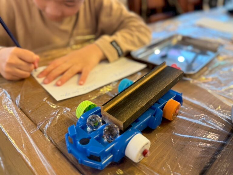 手前にできあがった車のような作品、こどもがその後ろで何か書いている