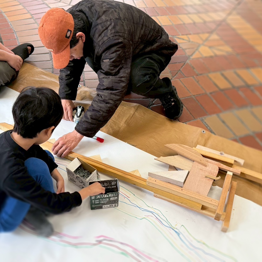男の子が木で恐竜を作っており、それを手伝うハブラボ講師ケンケン