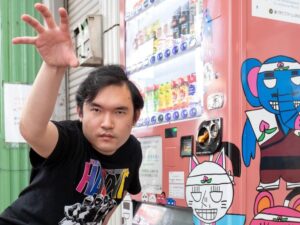 自身の作品がプリントされた自販機の前でポーズを決める堀本さん