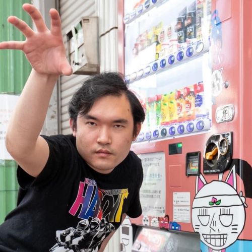 自身の作品がプリントされた自販機の前でポーズを決める堀本さん