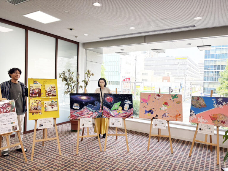 納品した作品が並んでいる横や後ろに立つアーティスト・miyaとアーティスト・小林智恵子