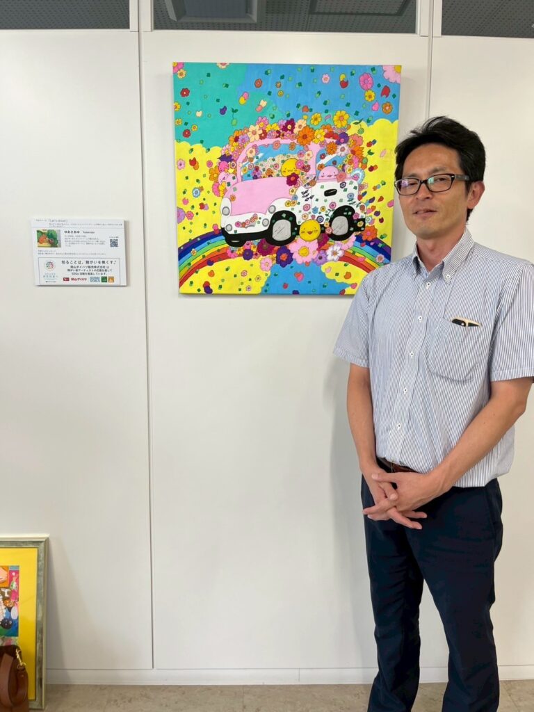 納品した作品（アーティスト・ゆあさあゆ作）の前に立って写る岡山ダイハツ中古車センターの方
