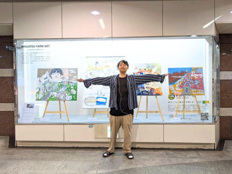 展示ケースの前で腕をひろげて写るアーティスト・Miya