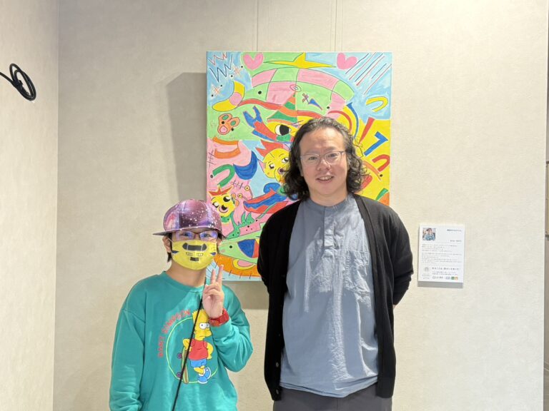 納品したアーティスト・KAITOの作品の前で笑顔で写るKAITOとひかり薬局の方