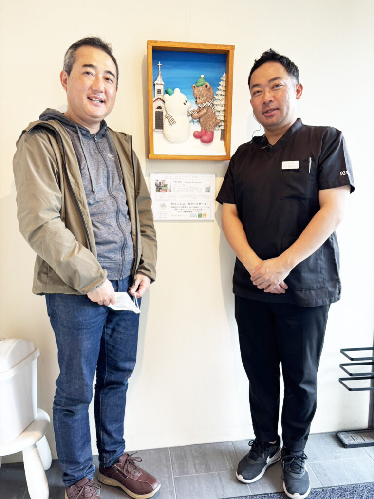 納品したアーティスト・下村良作 作「ともだちマフラー」（中央）と下村良作（左）きらり歯科院長（右）