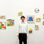 自分の作品のコーナー前で写るアーティスト・Miya(中央)斜め上を見ていてちょっと変顔