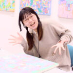 アーティスト・かなっぺ 机には制作中のキャンバス作品。はじけた笑顔で手を広げてポーズをとっている