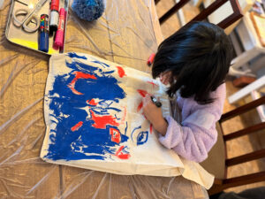 トートバッグに描いているようす、青井背景にところどころ赤で海の中を熱帯魚が泳いでいるように見える