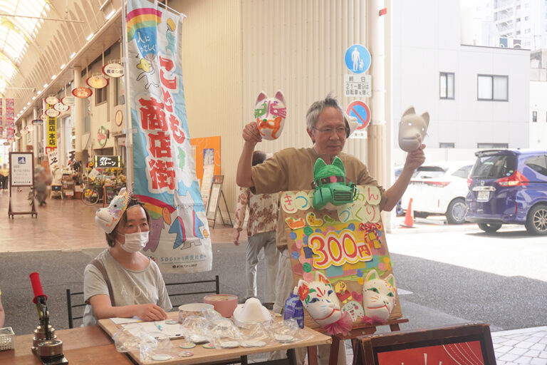 おめん絵付けブース、お面を両手にもってよびかけている